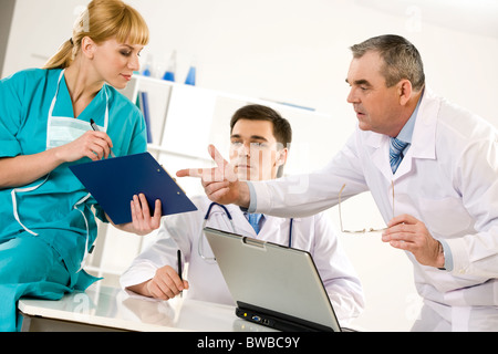 Photo of aged physician pointing at document in nurse’s hand with their colleague beteen them Stock Photo