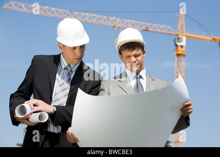 Portrait of two confident foremen looking at project on the background of sky and crane Stock Photo
