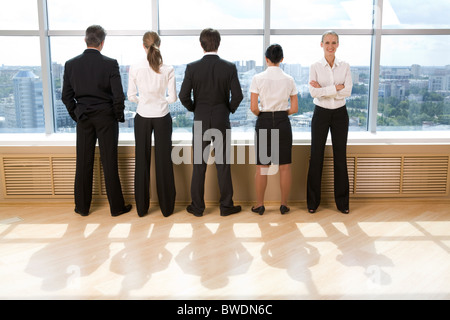 Rear view of business group standing in row and looking through window with their leader smiling at camera Stock Photo