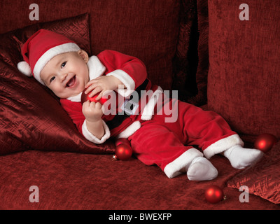License available at MaximImages.com - Happy smiling six month old baby boy in Santa Christmas costume lying on a sofa with a red bauble in his hands Stock Photo