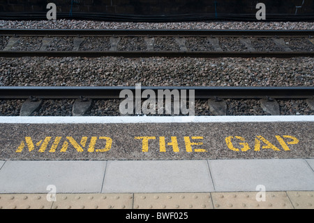 'Mind The Gap' stenciled lettering in yellow on the edge of a railway platform with the rail tracks in the background. Stock Photo