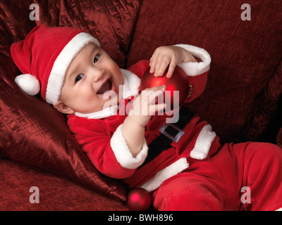 License available at MaximImages.com - Happy smiling six month old baby boy in Santa Christmas costume lying on a sofa with a red bauble in his hands Stock Photo