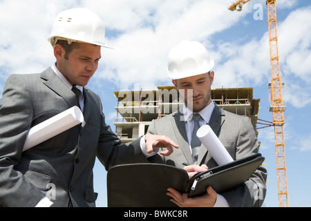 Portrait of two builders standing at building site and discussing new project held by one of them Stock Photo