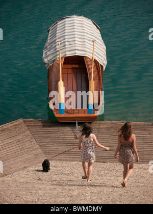 Two girls aged 14 and 16 with black spaniel run towards a moored gondola next to lake Bled Slovenia Stock Photo