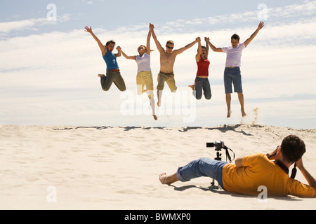 Professional photograpther taking shots of joyful friends jumping on sandy beach against blue sky Stock Photo
