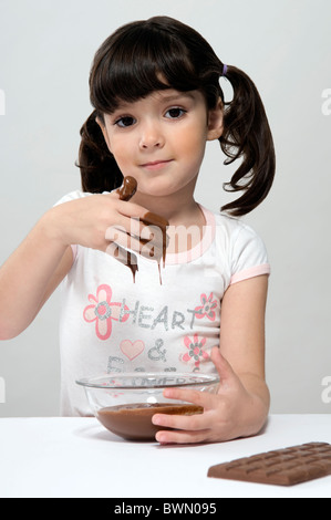 Girl looking at camera, putting her hand in a pot full of melted chocolate and a chocolate bar on the table Stock Photo