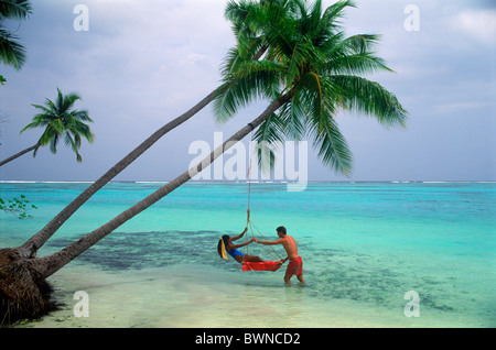 Couple under palm trees with woman sitting in swing on Meeru Island in the Maldives Stock Photo