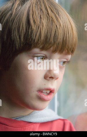 Eight year old boy with worried expression looking out of a window. Stock Photo