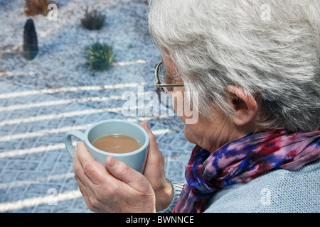 Everyday hygge scene with senior woman living alone wearing a scarf and holding a warm drink looking out of a window on a cold snowy day in winter. UK Stock Photo