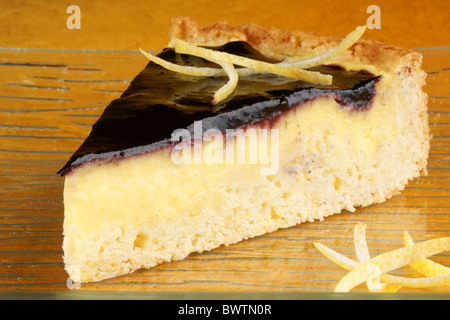 Slice of blueberry marmalade and custard cream tart served on a glass plate with lemon zest, over an orange background. Stock Photo