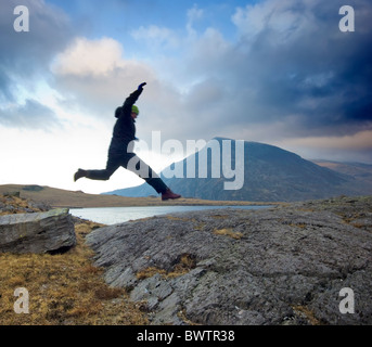 Hiker jumping across rocks Llyn Idwal, by footpath to Devils Kitchen, Snowdonia, North Wales. Pen yr Ole Wen  in the distance Stock Photo
