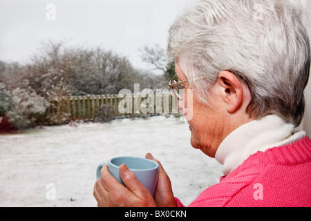 Elderly senior woman OAP lady living alone holding a hot drink and looking out through a window to snow in the garden. UK Stock Photo