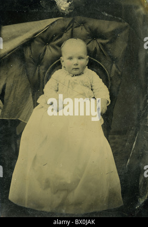 baby infant in portrait on black leather chair in photo studio white dress black and white tin type photograph strange off weird Stock Photo