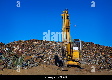 A large yellow crane works with a huge pile of scrap metal, getting it ready for recycling under clear blue skies. Stock Photo