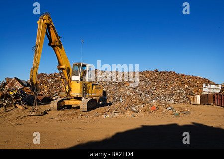 A large yellow crane works with a huge pile of scrap metal, getting it ready for recycling under clear blue skies. Stock Photo