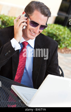 A happy smiling businessman wearing sunglasses and using a laptop computer and cell phone outside Stock Photo