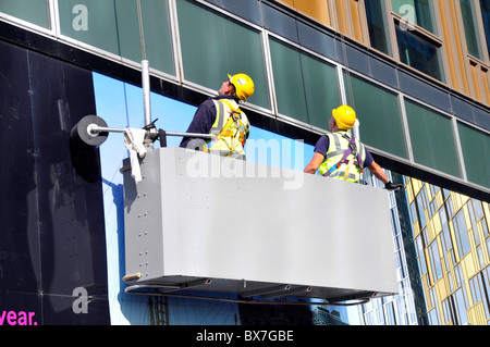 Two workmen in cradle supsended on cables inspecting the outside of a office block Stock Photo