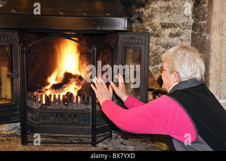 One elderly woman pensioner warming cold hands sat at a fireside in front of a roaring open log fire in a cosy warm living room. England UK Stock Photo