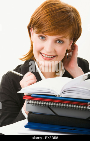 Portrait of cute red-haired woman looking from behind huge heap of documents and cases, holding pen and touching her head Stock Photo
