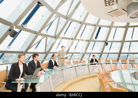 Businesspeople sitting at the round table listening to their colleague’s speech in large light conference hall with glassy walls Stock Photo