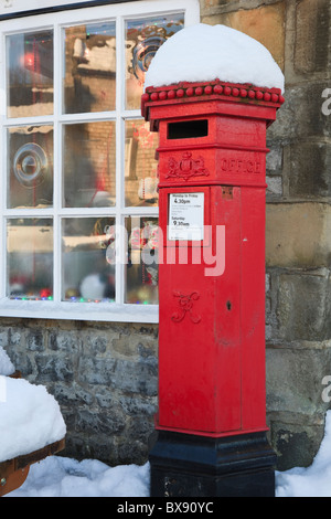 Old red Victorian postbox outside the post office with snow in winter. Hartington, Derbyshire, England, UK, Britain. Stock Photo