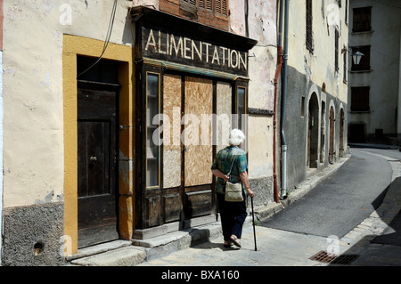 Old Woman with Walking Stick Walks Past a Boarded-Up & Closed Village Shop or General Store, Alimentation, Beauvezer, Alpes-de-Haute-Provence, France Stock Photo