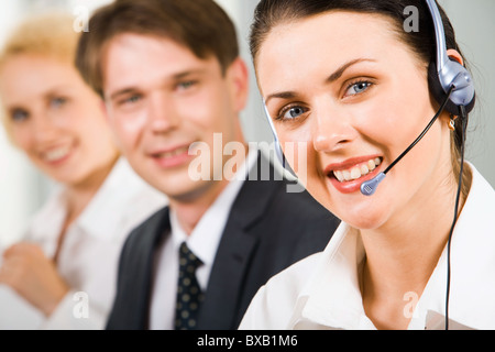 Portrait of smiling brunette businesswoman with blue headset two businesspeople in the background gazing at the camera Stock Photo