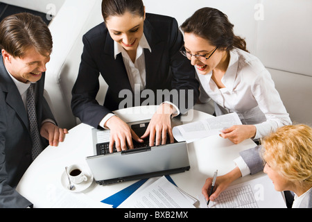 Three laughing businesswomen and a businessman sitting on the white sofas at the round table with opened laptop, documents and c Stock Photo