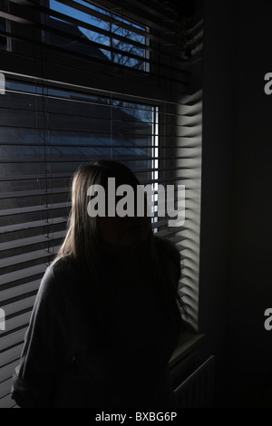 Woman sitting alone in the dark with her outline shown against a window blind. Stock Photo
