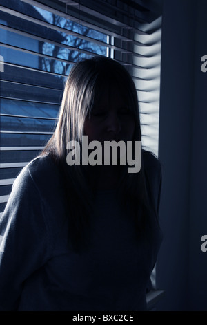 Portrait of an anonymous fair haired woman sitting with her back to a wooden window blind at night. Stock Photo