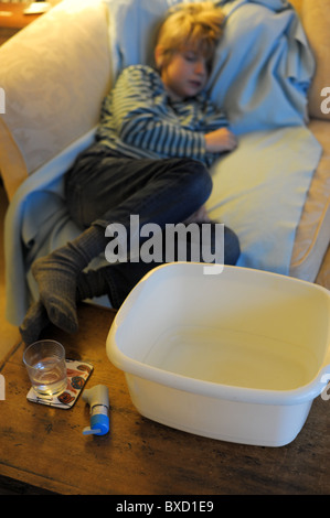 An ill child suffering from the winter vomiting bug asleep on a sofa with a sick bowl, glass of water and inhaler nearby Stock Photo