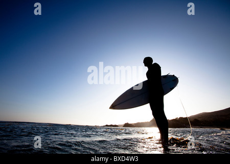 A male surfer stands on a rock while being silhouetted by the setting sun. Stock Photo