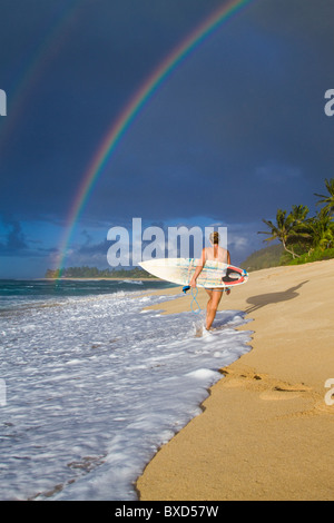 An amazing rainbow over Rocky Point, as a surfer girl walks along the beach, on the north shore of Oahu, Hawaii.