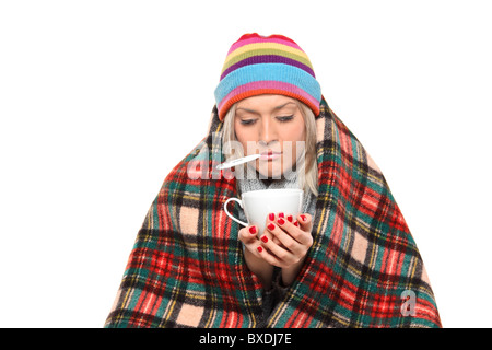 Ill woman covered with blanket holding a tea cup and a thermometer in her mouth Stock Photo
