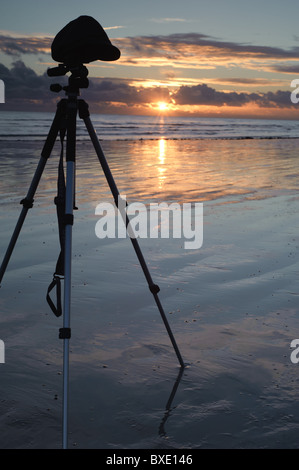 A photographers tripod stands on the wet beach in front of a sunset with clouds that is reflecting on the wet sand and water Stock Photo