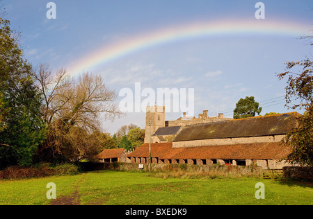 Rainbow over the church of St Mary in East Quantoxhead in Somerset with farm buildings bordering village green Stock Photo