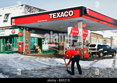 Man shovelling clearing snow from a Texaco petrol filling station forecourt after snowfall in bad winter weather. UK, Britain