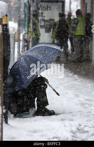 street beggar sitting under deloitte umbrella in the snow on a cold snowy winters day Belfast Northern Ireland Stock Photo
