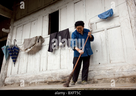 A Chinese woman sweeping the pavement or porch outside her rustic wooden house in a remote farmer's village in west China. Stock Photo