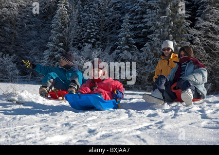 Three children sledding down a snowy hill while their mother looks on. Stock Photo