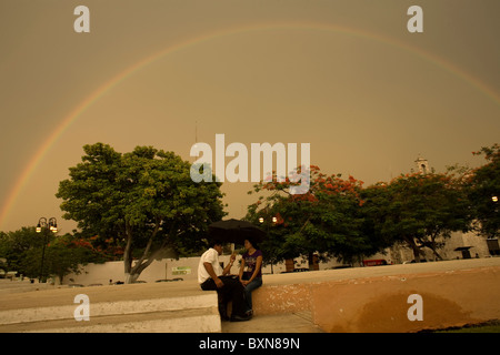 A couple sits in a public square under a rainbow using an umbrella as it rains in Yucatan state, Mexico, June 30, 2009. Stock Photo