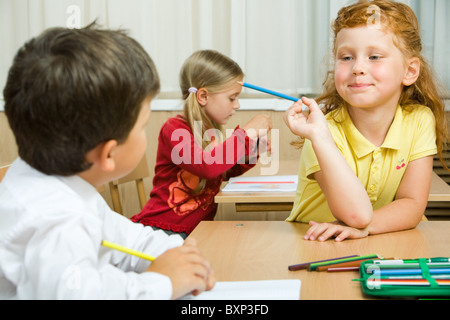 Photo of cute schoolgirl with blue pencil in hand looking at her classmate during lesson Stock Photo