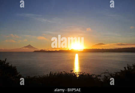 Yellow orb of the rising sun shining on Lake Llanquihue, between the Osorno and Calbuco Volcanoes, seen from Puerto Varas, Chile Stock Photo