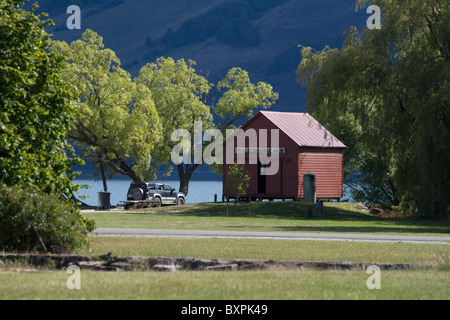 A red/brown wooden hut stands by the pier in the middle of Glenorchy at the head of Lake Wakatipu, with  'Glenorchy' on it