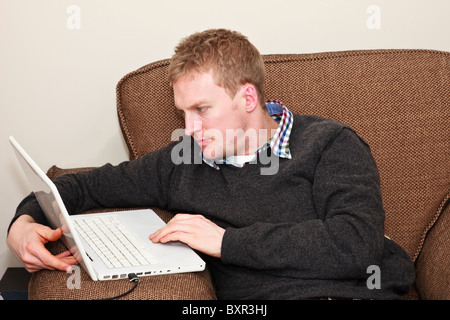 Young Millennial man looking at Facebook using a laptop computer resting on arm of an easy chair at home. England, UK, Britain. Stock Photo