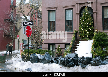 Bags of trash sit on a pile of snow awaiting pick-up in front of a brownstone decorated for Christmas Stock Photo