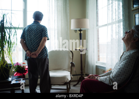 Old woman sits with head tilted back while old man stands looking out window. Stock Photo