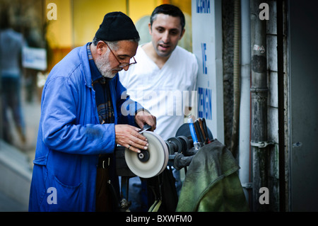 Knife Sharpener Street Vendor Istanbul Turkey / ISTANBUL, Turkey — A street vendor uses a mobile grinder to sharpen knives in downtown Istanbul. The older craftsman, wearing a blue work coat, intently focuses on his task while a younger man observes in the background. This traditional mobile service provides essential knife maintenance for the local community. Stock Photo
