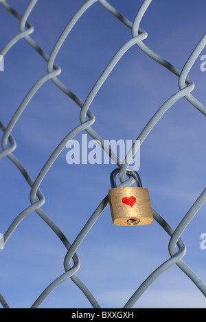 Locked padlock with heart hanging on a chain linked fence with a blue sky in the background. Stock Photo