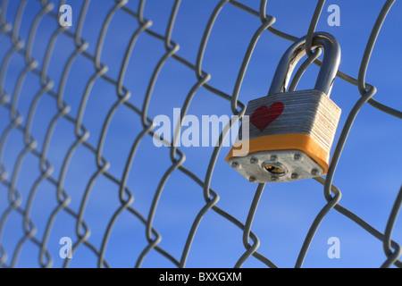 Locked padlock with heart hanging on a chain linked fence with a blue sky in the background. Stock Photo